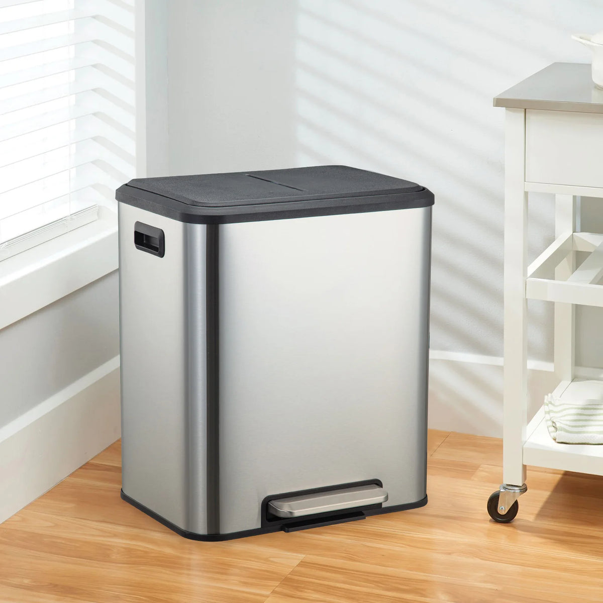 A stainless steel dual compartment step trash can with a black soft-close lid sitting on a wooden floor.