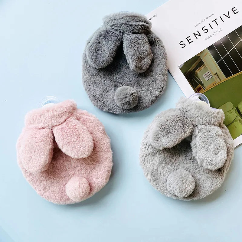 Three plush rabbit hot water bottles in shades of gray and pink resting on a light blue surface.