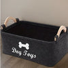 A gray felt pet toy storage basket with wooden handles sits on a wooden floor.
