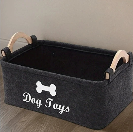 A gray felt pet toy storage basket with wooden handles sits on a wooden floor.
