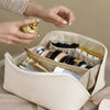 A hand is opening a beige leather travel makeup organizer bag revealing various cosmetic and jewelry items inside.