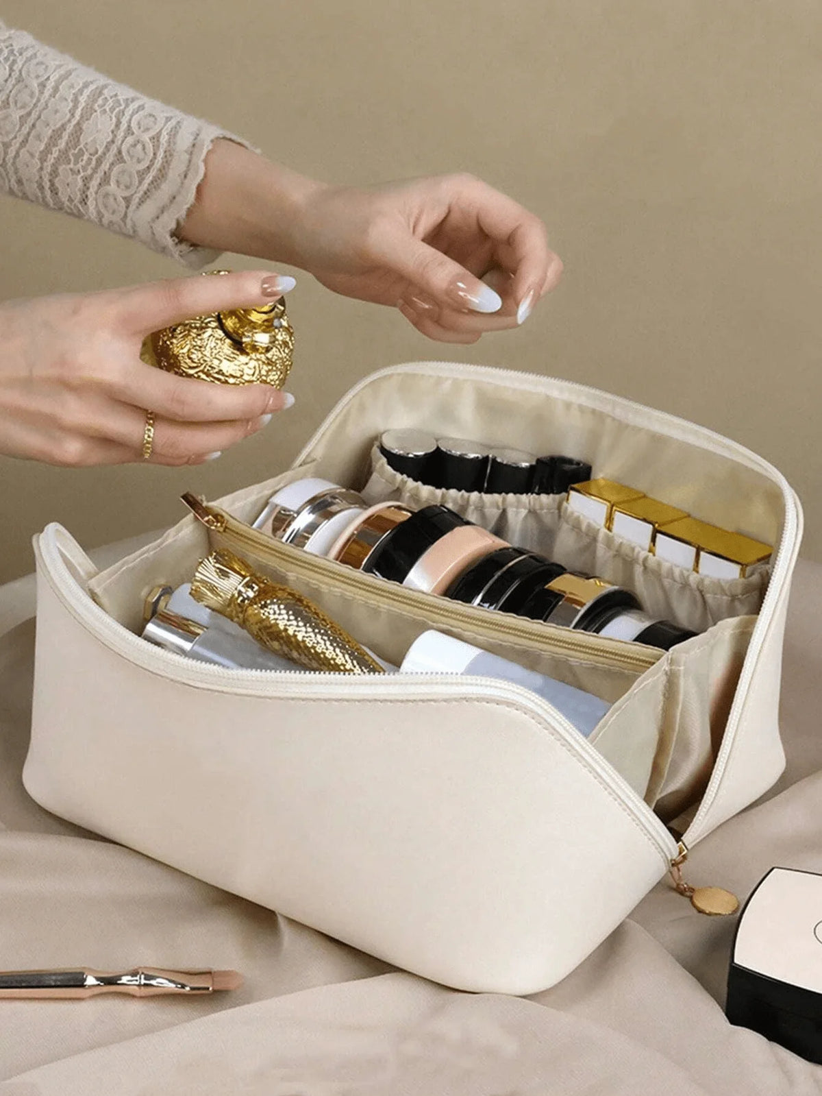 A hand is opening a beige leather travel makeup organizer bag revealing various cosmetic and jewelry items inside.
