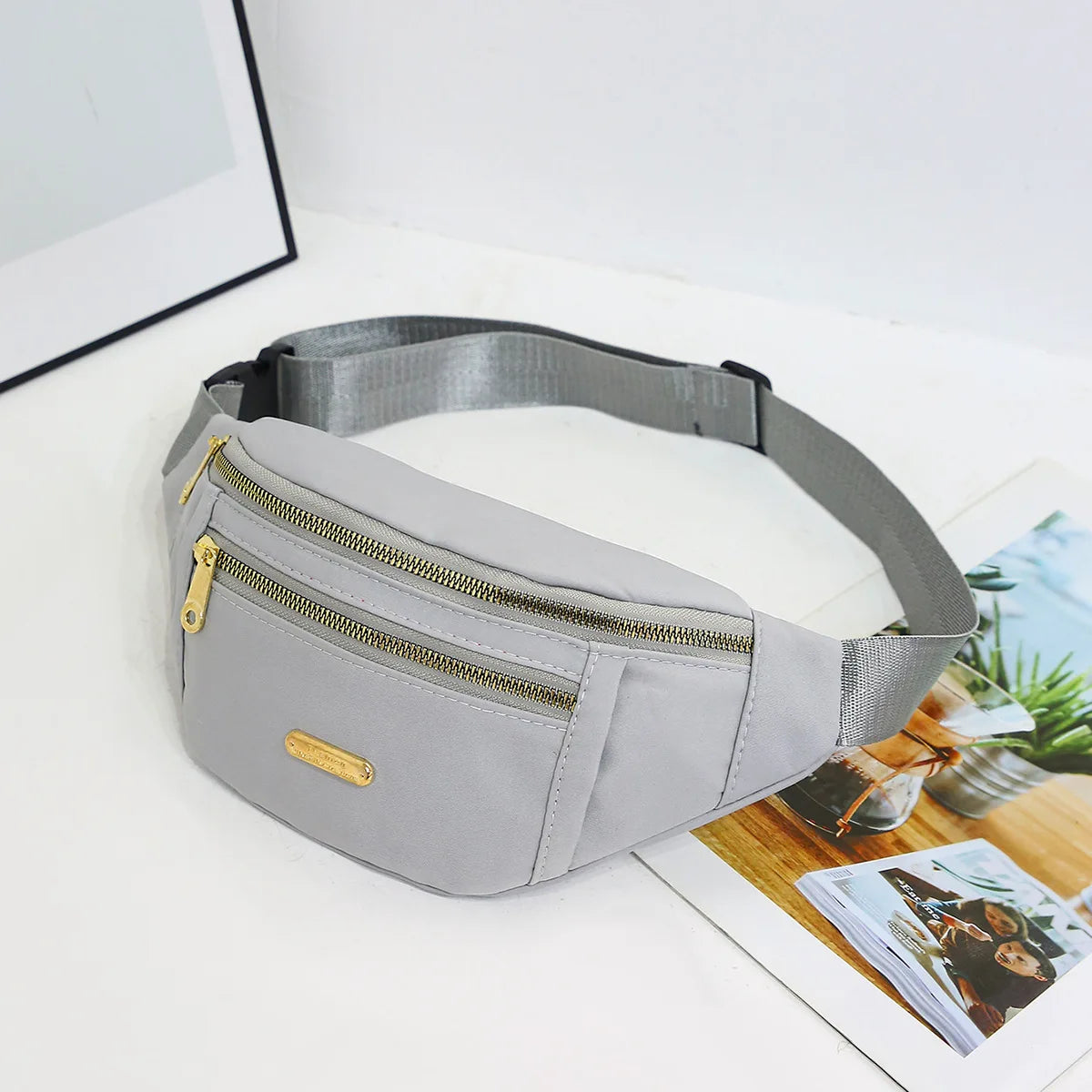 A light gray nylon waist pack with a zipper and gold accent is displayed on a white surface.