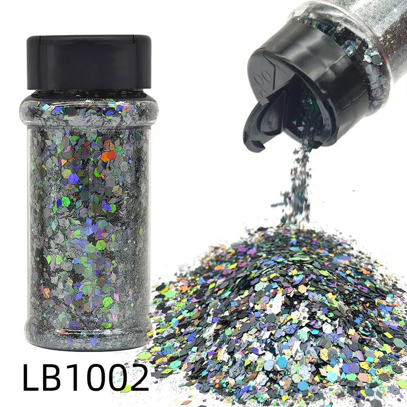 A glass jar of holographic chunky nail glitter mix with a black shaker top next to a pile of loose glitter.
