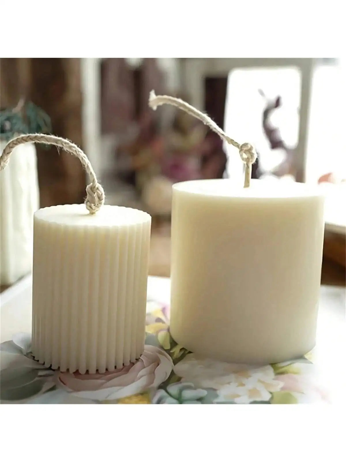 Two white pillar candles with natural twine wicks sit on a floral surface.