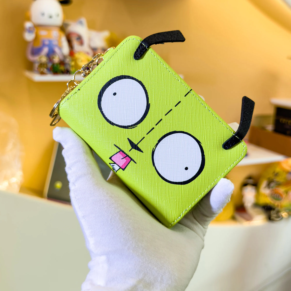 A bright lime green cartoon print compact wallet with large black eyes and a stitched line detail is held by a hand.
