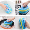 Hands demonstrate the use of a blue and yellow multipurpose sponge cleaning brush for cleaning various surfaces.