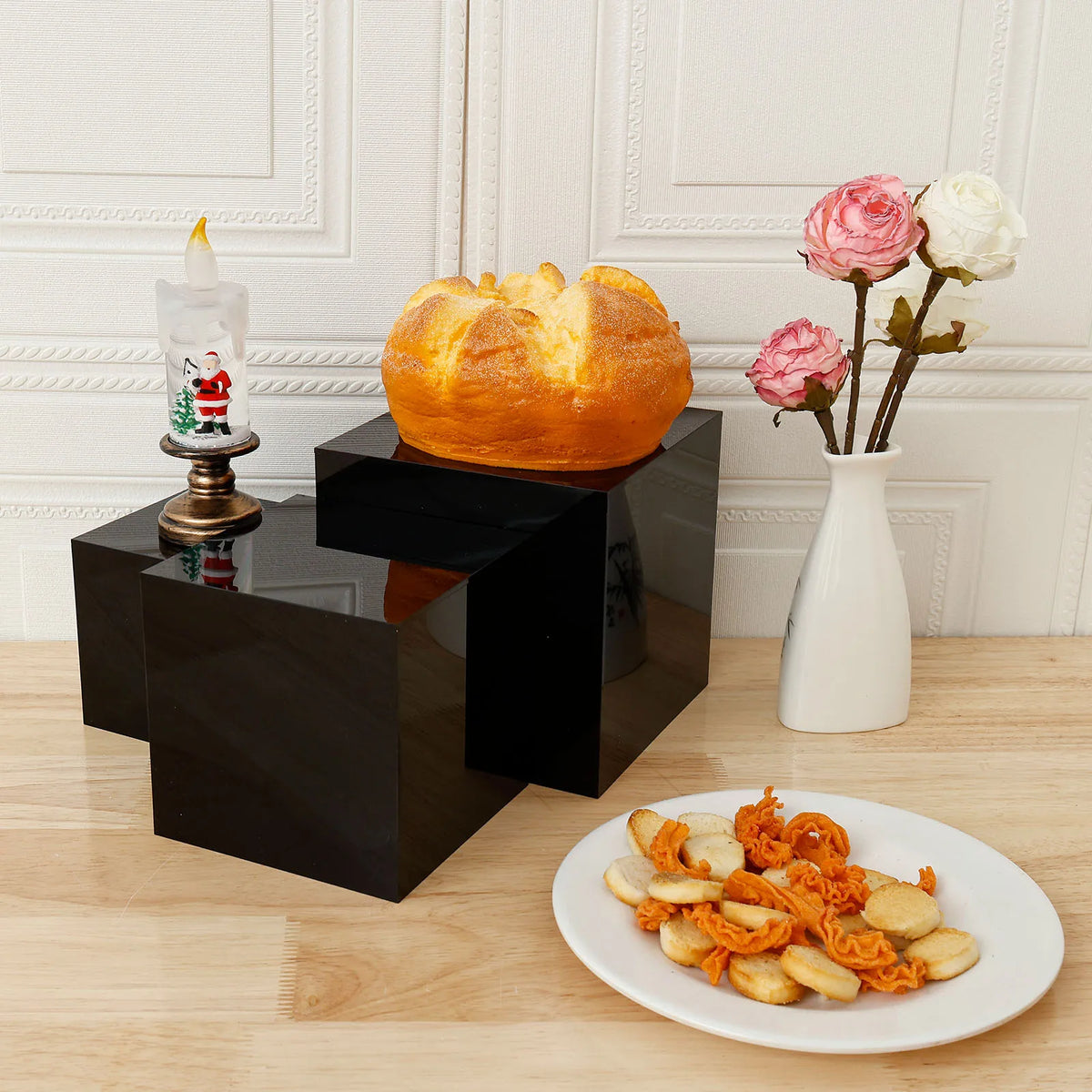 A golden baked loaf of bread sits on a black acrylic display riser next to a white candle and pink roses.