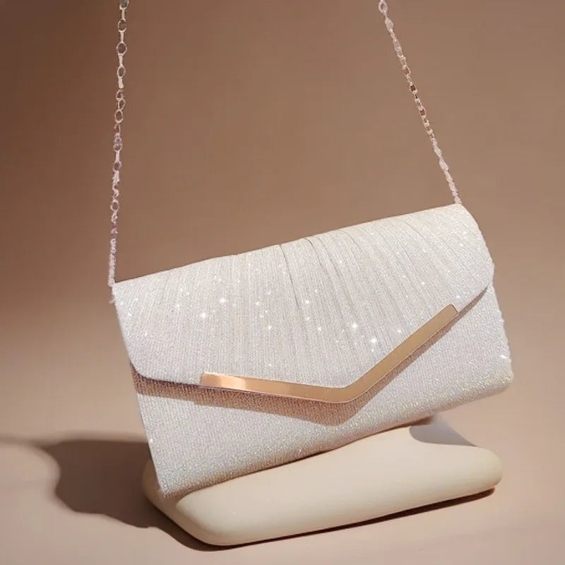 A white pleated glitter envelope clutch with a gold accent and a detachable chain strap resting on a light surface.
