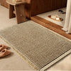 A beige waffle weave bath mat with tassels placed on a wooden surface next to a cabinet.