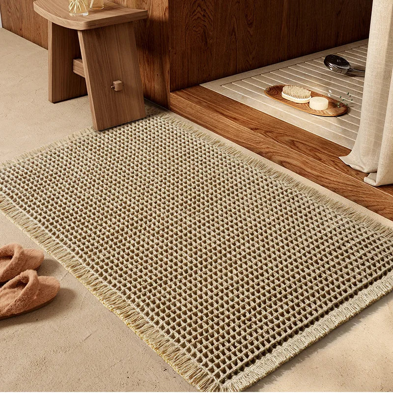 A beige waffle weave bath mat with tassels placed on a wooden surface next to a cabinet.