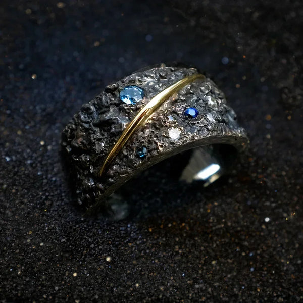 Anillo de chapado de plata con una piedra oscura texturizada y pequeños diamantes azules, montado en banda de oro.