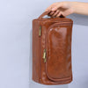 A brown leather toiletry bag with a zipper closure is being held by a hand against a light gray background.