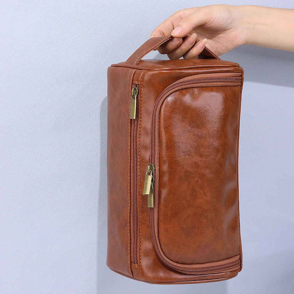 A brown leather toiletry bag with a zipper closure is being held by a hand against a light gray background.