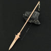 A slender, rose gold metal sword keychain with a decorative blade rests on a dark stone.