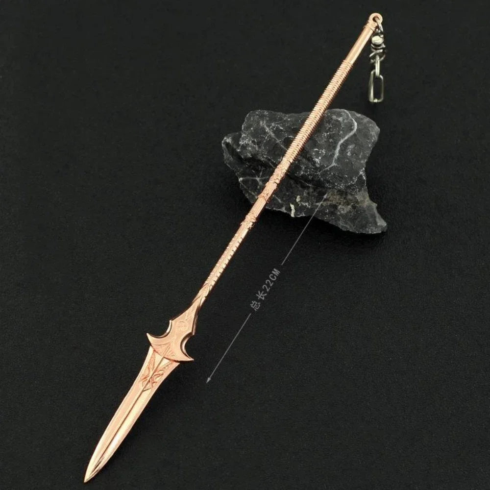 A slender, rose gold metal sword keychain with a decorative blade rests on a dark stone.