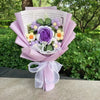 Handgemachter Häkelblumenstrauß mit lila und gelben Blumen, in rosa Papier eingewickelt und mit weißer Schleife gebunden.