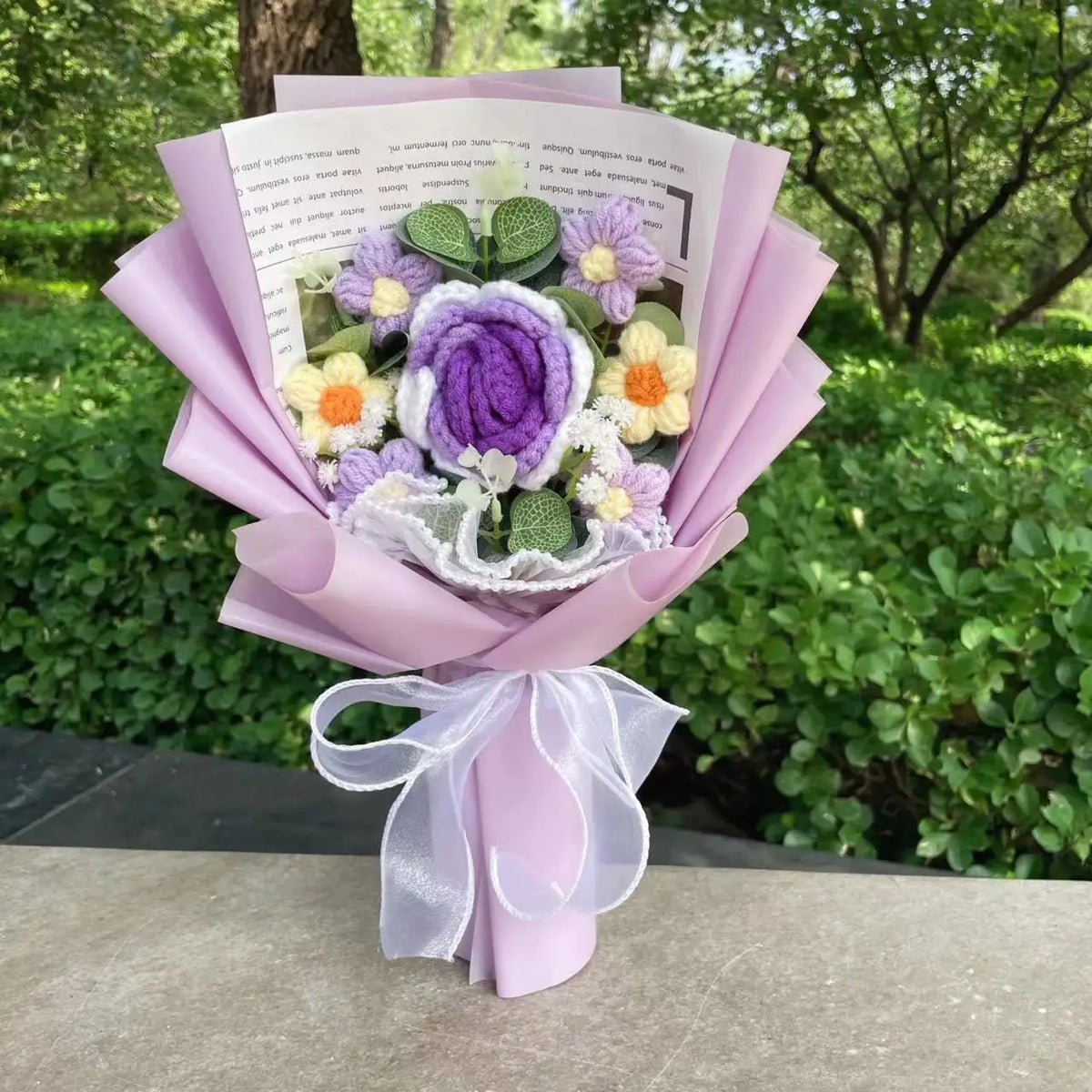Handgemachter Häkelblumenstrauß mit lila und gelben Blumen, in rosa Papier eingewickelt und mit weißer Schleife gebunden.