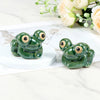 Hand-painted ceramic frog toilet bolt covers resting on a white surface with soft floral greenery in the background.