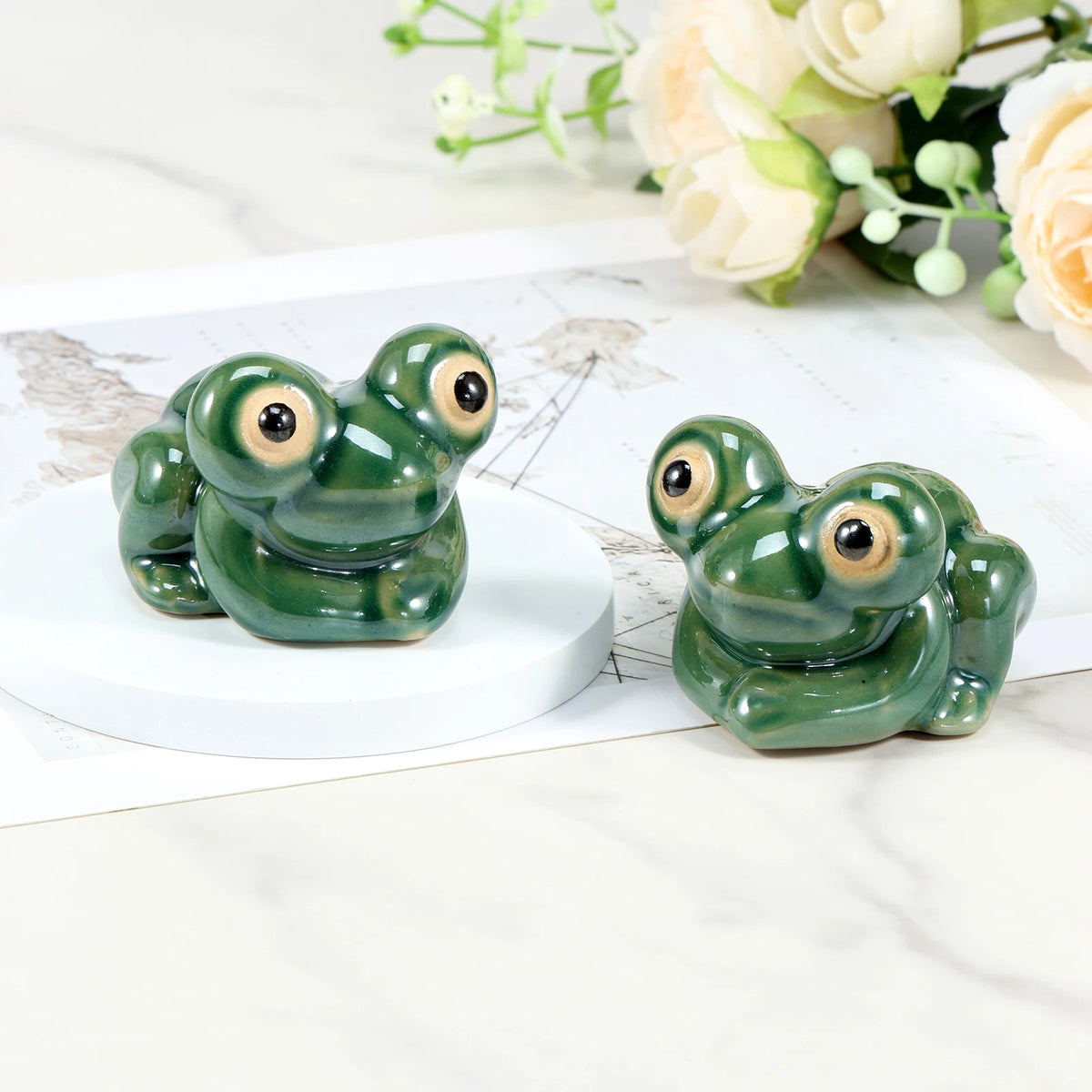 Hand-painted ceramic frog toilet bolt covers resting on a white surface with soft floral greenery in the background.