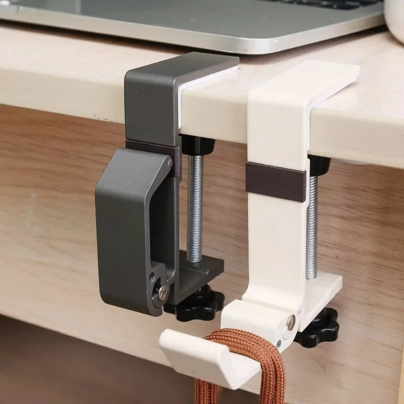 A foldable aluminum desk hook with a gray clamp and white arms is mounted on a wooden desk surface.