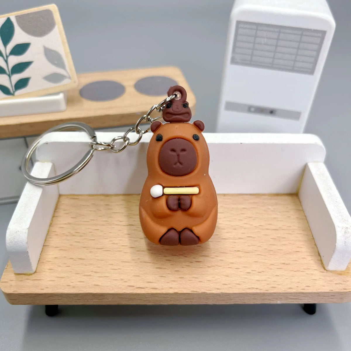 An adorable capybara keychain charm with a gold chain resting on a light wooden tray.
