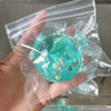 A hand holds a clear plastic bag containing teal-colored ocean squishy stress balls and small sea shells.
