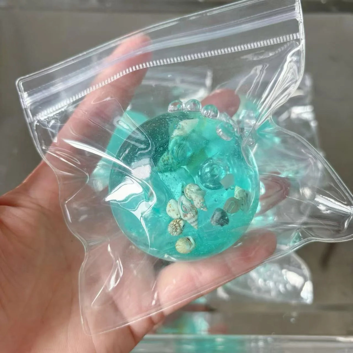 A hand holds a clear plastic bag containing teal-colored ocean squishy stress balls and small sea shells.