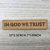 Brown woven patch with black text reading IN GOD WE TRUST on a light wooden surface.