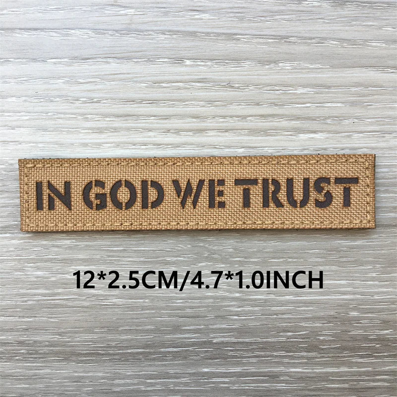 Brown woven patch with black text reading IN GOD WE TRUST on a light wooden surface.