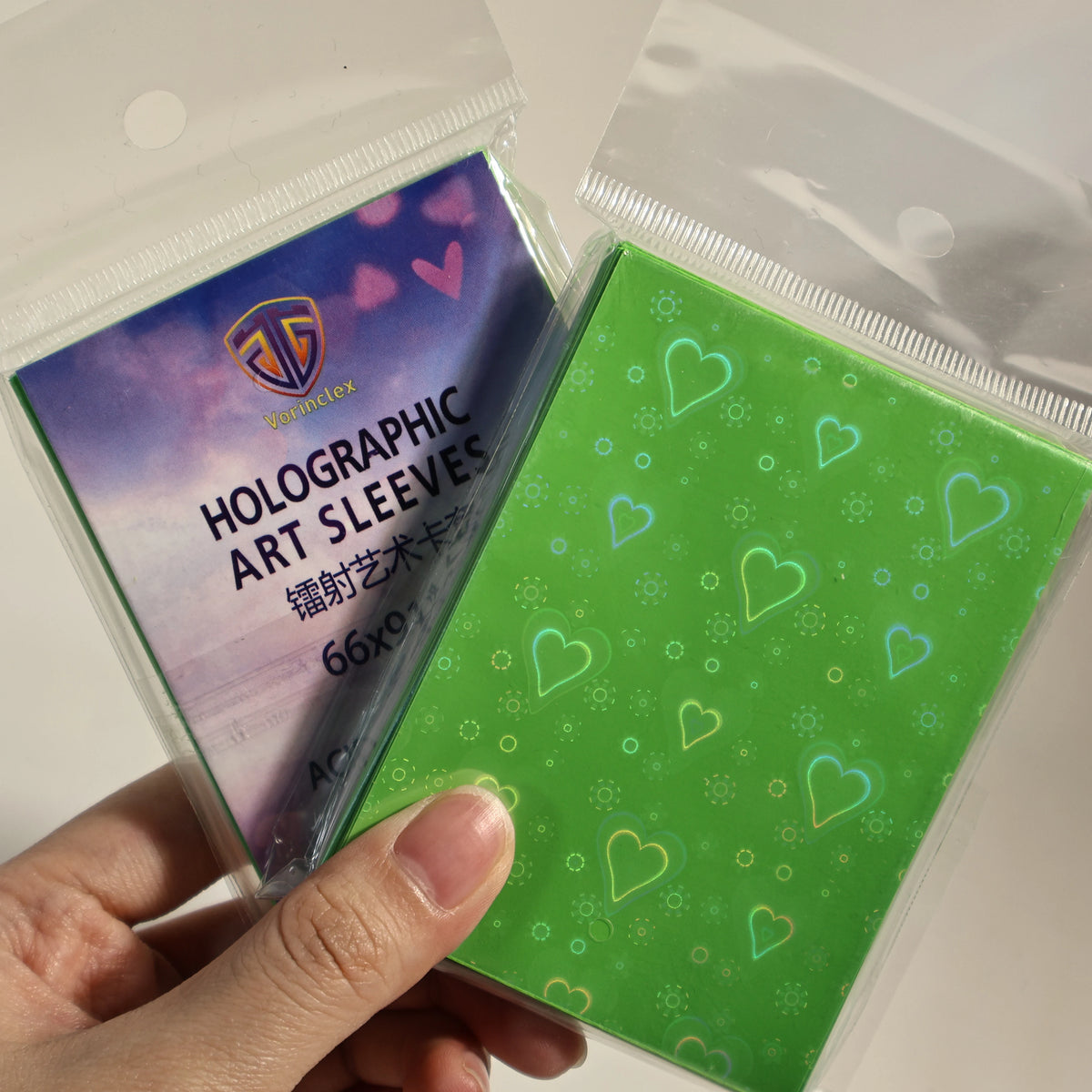 A hand holds a pack of holographic art sleeves with heart shapes inside clear plastic packaging.