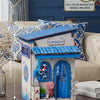 A wooden beach corner DIY book nook kit featuring a blue door and window with LED lighting displayed on a sofa.