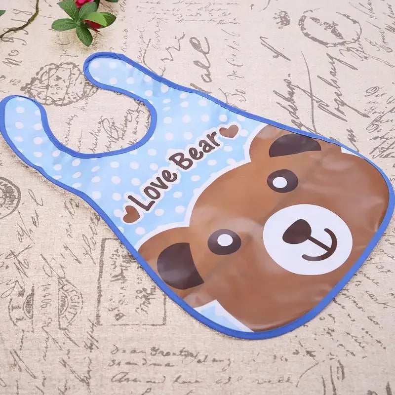 A blue and brown wipeable baby bib featuring a bear design with a catch-all pocket for weaning toddlers.