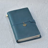 A dark teal leather refillable journal with a gold heart pendant resting on a light gray surface.