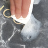 A hand is holding a white quick-dry diatomite bath mat with a non-slip backing on a dark gray surface.