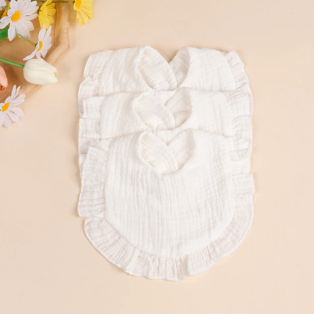 A set of three white soft cotton ruffle bibs laid on a light background with yellow flowers.