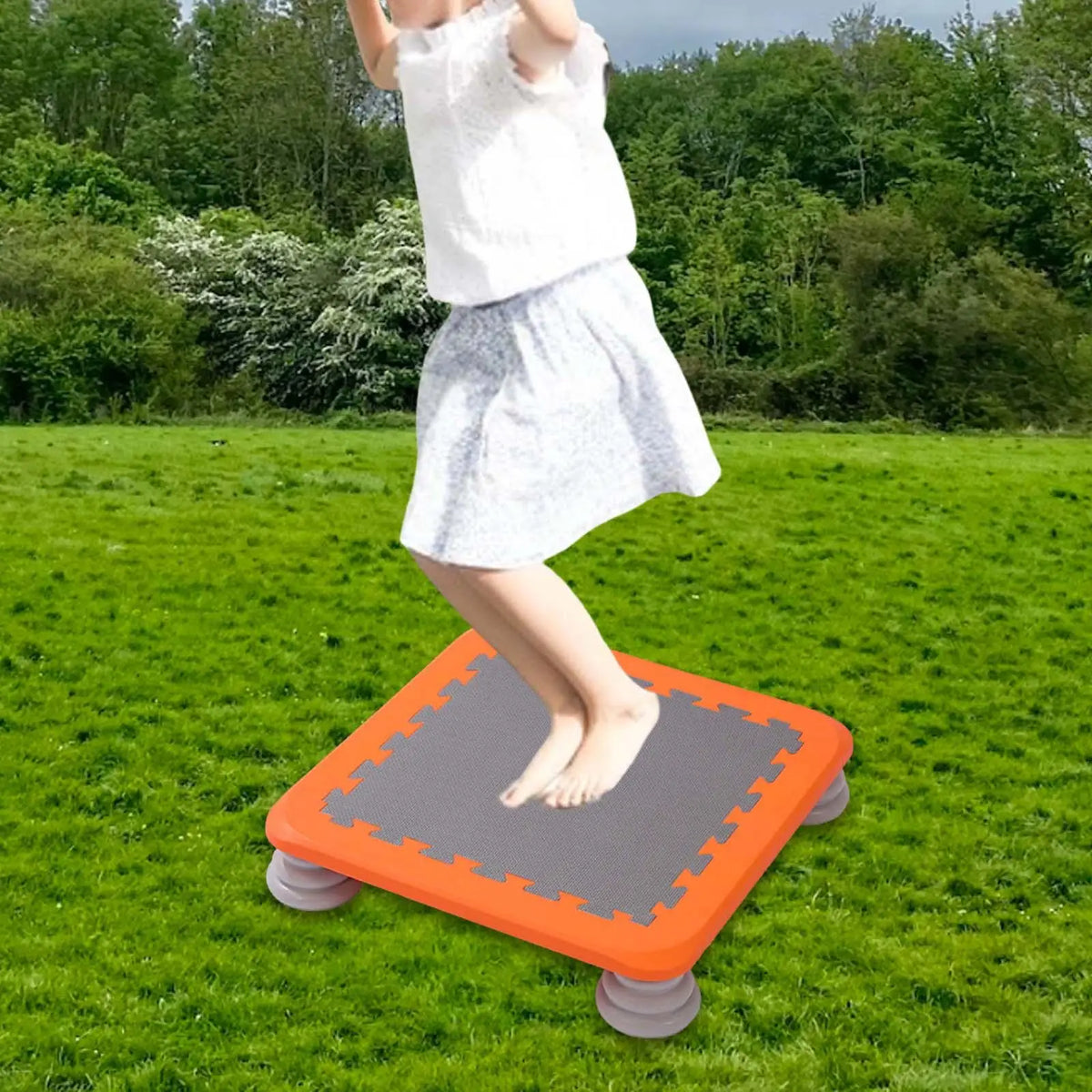 A child is standing on an orange mini trampoline with a gray mat in a grassy field.