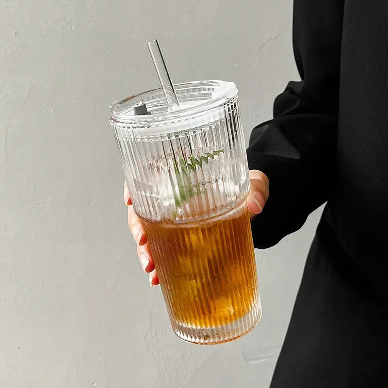 Une main tient un verre à pilons rayé de 600ml avec couvercle et paille contenant une boisson de couleur ambrée.
