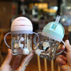 A hand holds a clear plastic straw cup with handles, featuring a white elephant design, against a blurred background.