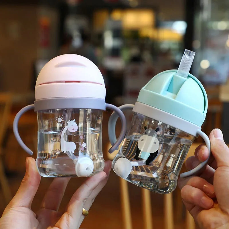 A hand holds a clear plastic straw cup with handles, featuring a white elephant design, against a blurred background.