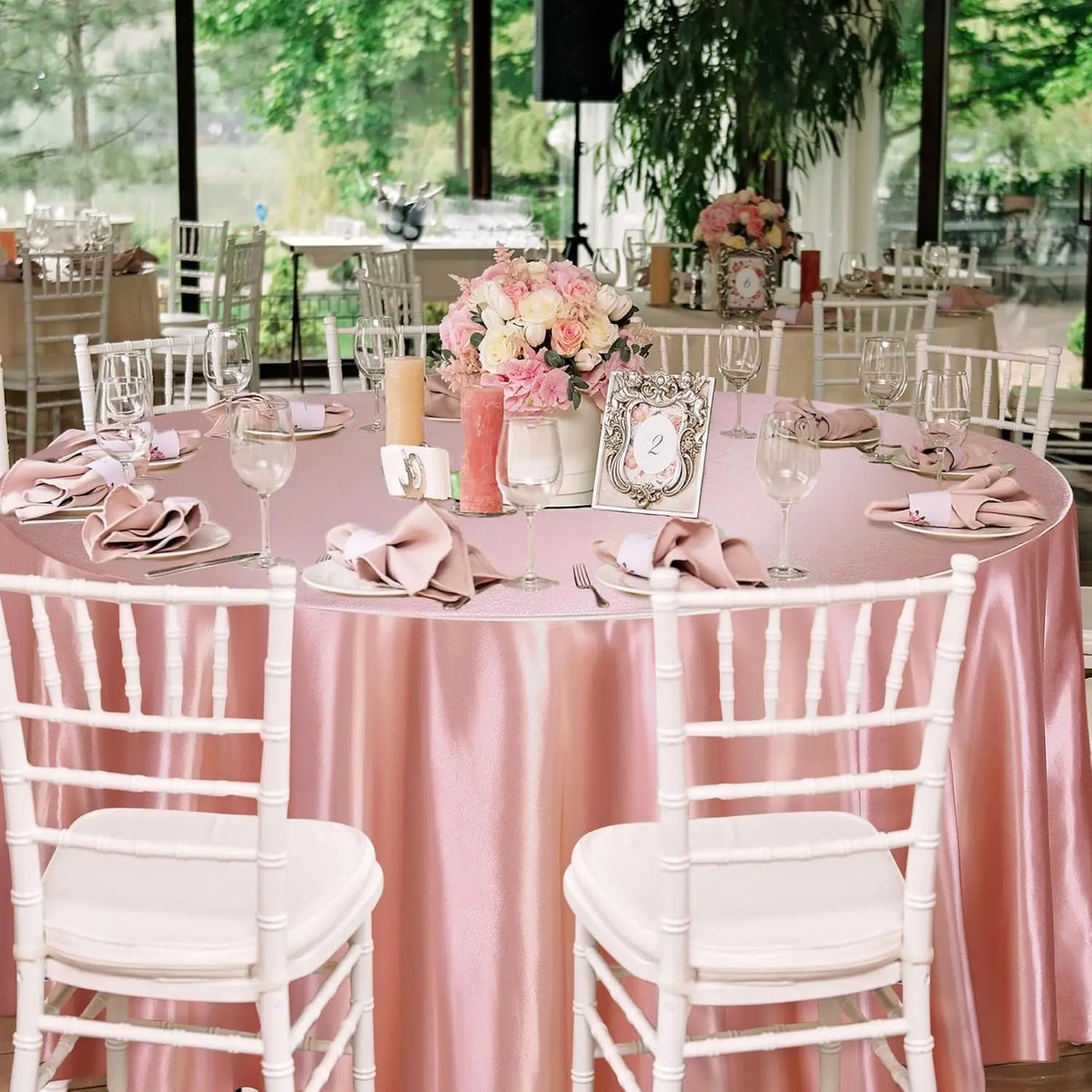 A round table set for an event featuring a pink satin tablecloth, white chairs, and floral centerpieces.