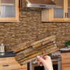 A hand holds a rectangular piece of wood tile stickers against a textured stone backsplash in a kitchen setting.