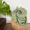 A rolled green linen fitted sheet with a deep pocket is placed inside a woven basket next to a large green monstera leaf.