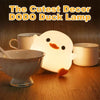 A glowing white DODO duck lamp sits next to two mugs on a wooden surface.