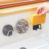 A hand is holding a yellow sponge over a stainless steel suction cup holder mounted on a white sink surface.