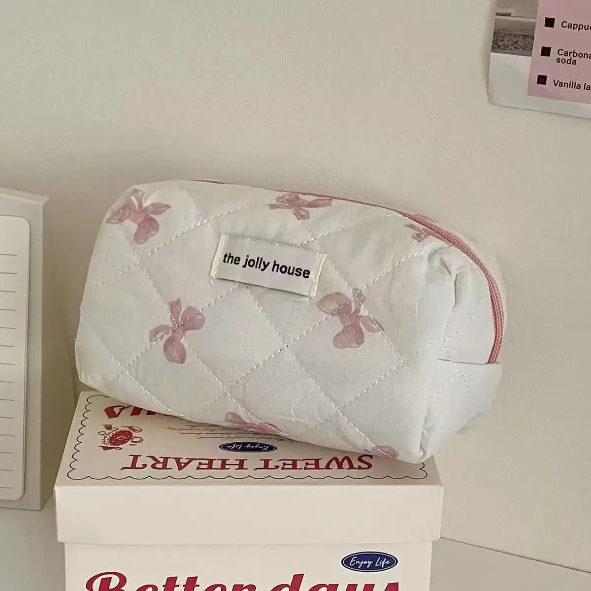 A white quilted cosmetic bag with pink bow print resting on a box.