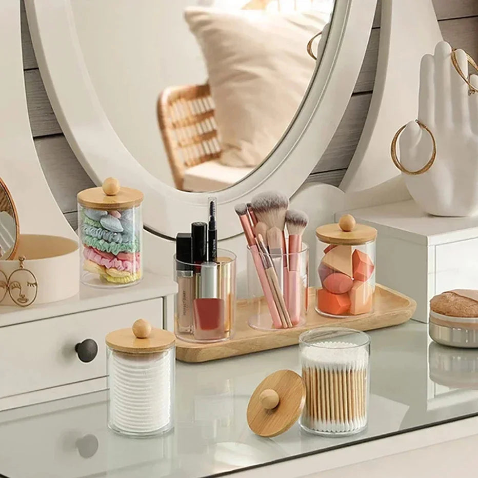 Clear acrylic apothecary jars with wooden lids and various beauty tools displayed on a white vanity surface.