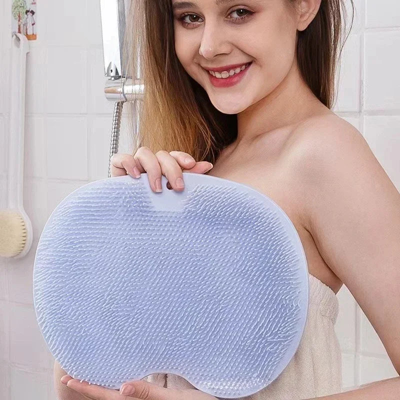 A woman holds a light blue silicone shower mat with exfoliating bristles in front of a bathroom mirror.