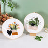 A circular embroidery kit featuring pre-printed designs of a cat and cactus in a floral pot on a white background.
