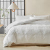 White duvet cover set with delicate embroidered leaf patterns on a bed against a light wall and a black grid background.